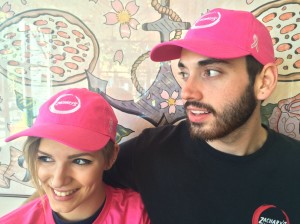 Two people wearing bright pink caps and black shirts with logos. They are smiling, standing against a wall with a floral design. The cap logo includes text, and theres a small pink ribbon symbol on the cap.