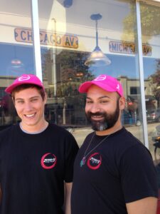 Two men wearing matching bright pink caps and black shirts with a logo on the chest are smiling. Theyre standing in front of a building with large windows, displaying street signs labeled Chicago Av and Michigan Av.