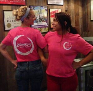 Two people stand back to back, smiling at each other. Theyre wearing pink T-shirts with Zacharys Chicago Pizza logos. The background features a wooden wall with framed posters and awards.