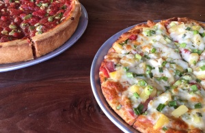 Two pizzas on a wooden table: one is a deep-dish with pepperoni and green peppers, partially visible on the left; the other is a thin crust topped with pineapple, ham, and green onions on the right.