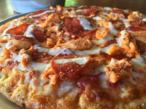 Close-up of a pizza topped with pepperoni, chunks of grilled chicken, and melted cheese. The crust appears golden and crispy, with a sprinkling of herbs. The pizza is on a plate on a wooden table.