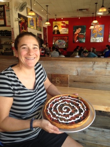 A person smiling holds a deep-dish pizza with a spiral topping, sitting at a wooden table in a lively restaurant. The walls are adorned with colorful posters, and people are dining in the background.