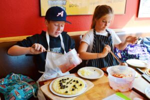 Two children are making pizzas at a table. The boy, wearing a baseball cap, spreads sauce on dough, while the girl sprinkles toppings. Both are in aprons. Various containers of ingredients are on the table. Bright, colorful walls are in the background.