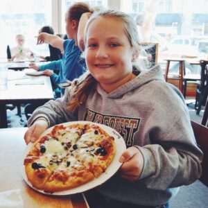 A smiling girl in a gray hoodie holds a plate with a small pizza topped with olives and cheese. She is sitting at a table in a restaurant with other people in the background.