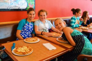 Three smiling girls sit at a table with personal-sized pizzas in front of them. They are wearing aprons and appear to be in a restaurant setting with a red wall and framed artwork in the background.