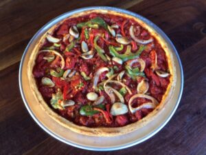 A deep-dish pizza with a thick crust, topped with tomato sauce, sliced onions, green bell peppers, and pieces of tomato, served on a metal platter on a wooden table.