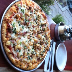 A pizza topped with cheese, diced potatoes, bacon, and green onions on a round metal tray. It sits on a wooden table with a white plate, fork, knife, and shakers of parmesan and chili flakes nearby. Garden view outside the window.