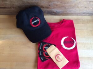 Black cap and red T-shirt with a Zacharys Chicago Pizza logo on a wooden surface. Nearby are three black gift cards and a small brown envelope with the same logo.