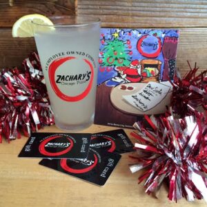 A glass with a lemon slice and the Zacharys Chicago Pizza logo is surrounded by red tinsel and gift cards. In the background, a colorful painted image depicts a holiday scene with a tree and presents.