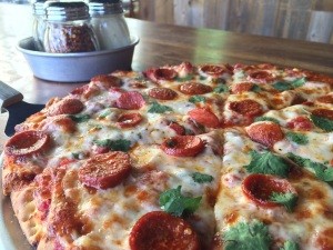 Close-up of a pepperoni pizza with melted cheese and herbs on a wooden table. In the background, there are small jars containing red pepper flakes and grated cheese.