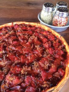 Close-up of a deep-dish pizza topped with sausage and pepperoni, with a crispy crust. Next to the pizza is a round tray holding shakers of oregano, crushed red pepper, and Parmesan cheese. The setting is a wooden table.