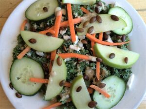 A fresh salad with kale, sliced cucumber, green apple wedges, and carrot sticks, topped with pumpkin seeds and drizzled with a creamy dressing, served on a white plate.