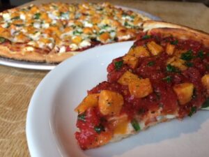 Close-up of a slice of pizza with a thick tomato sauce, chunks of butternut squash, and herbs, on a white plate. In the background, a whole pizza with various toppings, including cheese and green herbs, sits on a wooden table.