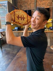 A man holds a football-shaped pizza with pepperoni laces near his head, mimicking a throwing pose. He is in a restaurant with a brick wall and menu board in the background.