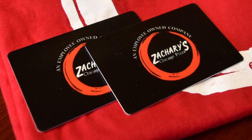 Two black Zacharys Chicago Pizza gift cards rest on a red and white fabric. Each card features a circular red design and the text An Employee Owned Company above the logo.