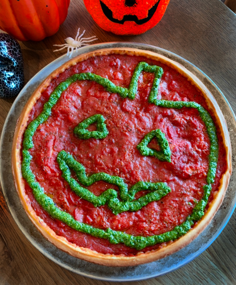 A pizza with a jack-o-lantern face, made with green pesto on a red tomato sauce base. It sits on a wooden table, surrounded by Halloween decorations including a red pumpkin lamp and a white spider figurine.