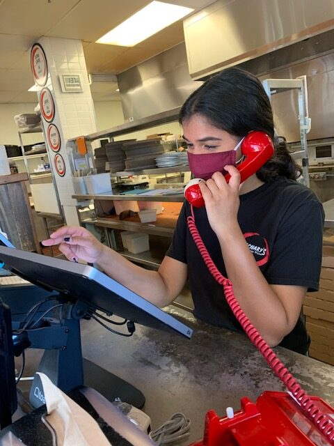 A person wearing a black shirt and face mask is speaking on a red corded phone while touching a screen at a counter in a restaurant kitchen. Behind them, there are shelves with plates and other kitchen equipment.