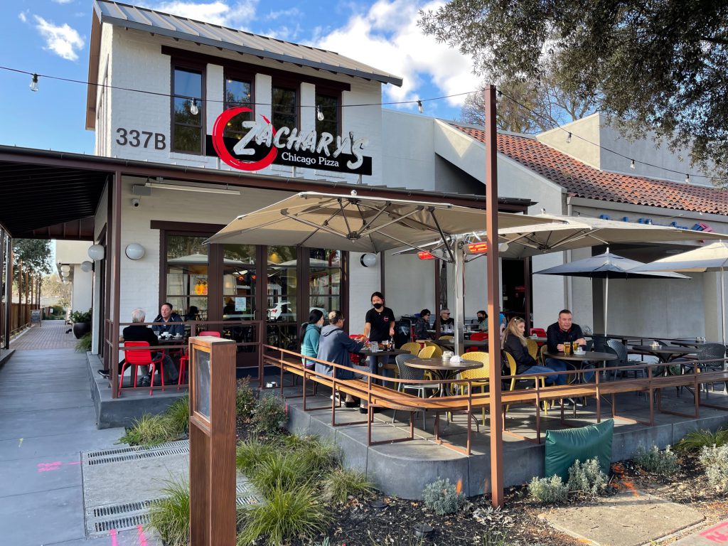 Outdoor seating area of Zacharys Chicago Pizza on a sunny day. Several people are dining at tables under large umbrellas. The building features a modern design with a combination of wood and white materials. The address 337B is visible.