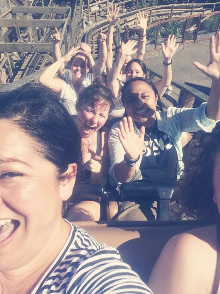 A group of smiling people, seated in a roller coaster car, raise their hands in excitement. The wooden tracks curve behind them under a clear blue sky. Everyone appears to be joyful and enjoying the ride.