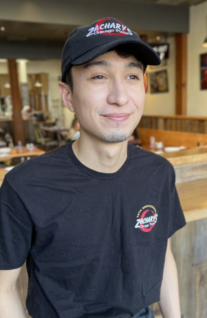 A person wearing a black cap and T-shirt with Zacharys logo stands in a restaurant. They have a slight smile and the background shows wooden furnishings and blurred diners.