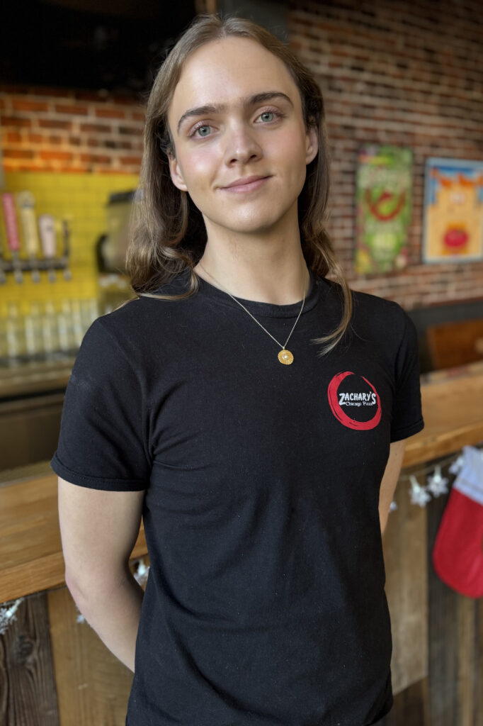 Person with long hair wearing a black Zacharys T-shirt, standing in a restaurant with a brick wall and a bar in the background. They are smiling slightly, with hands behind their back.