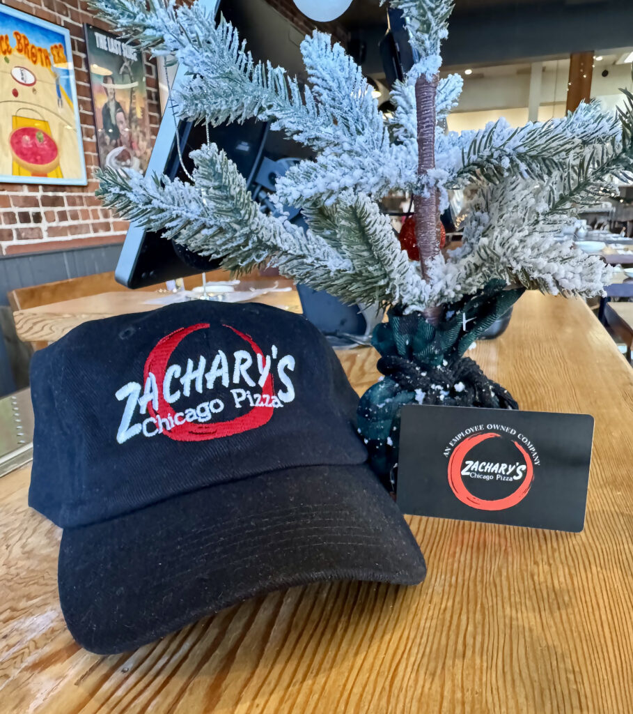 A black cap with Zacharys Chicago Pizza logo sits on a wooden table next to a frosted Christmas tree decoration and a gift card. The setting is indoors with a rustic, festive ambiance.