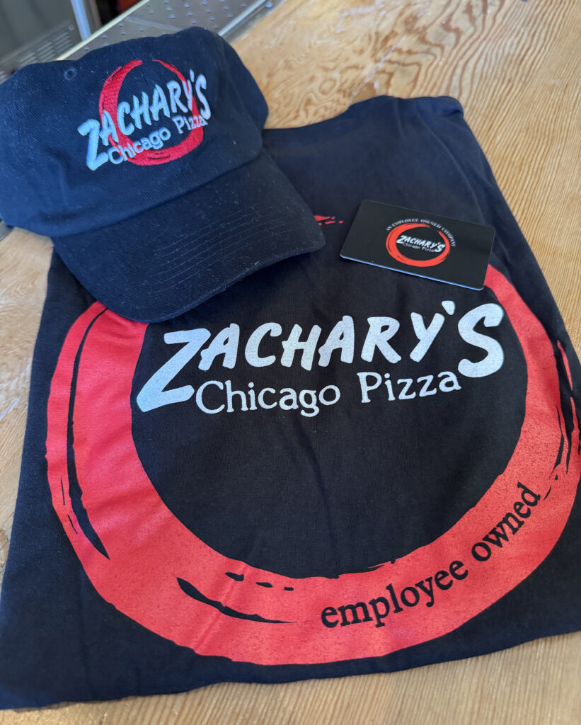 A navy blue cap and black t-shirt featuring the Zacharys Chicago Pizza logo are placed on a wooden surface. The t-shirt includes the phrase employee owned. A black card with the same logo is next to them.