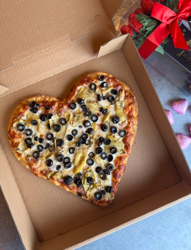 Heart-shaped pizza topped with olives and artichokes in an open cardboard box. Next to it is a bouquet of strawberries with a red ribbon.