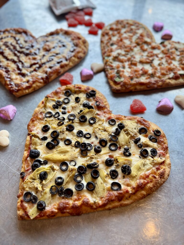 Three heart-shaped pizzas with various toppings, like black olives and artichokes, are set on a metal surface. Scattered candy hearts surround the pizzas, adding a festive touch.