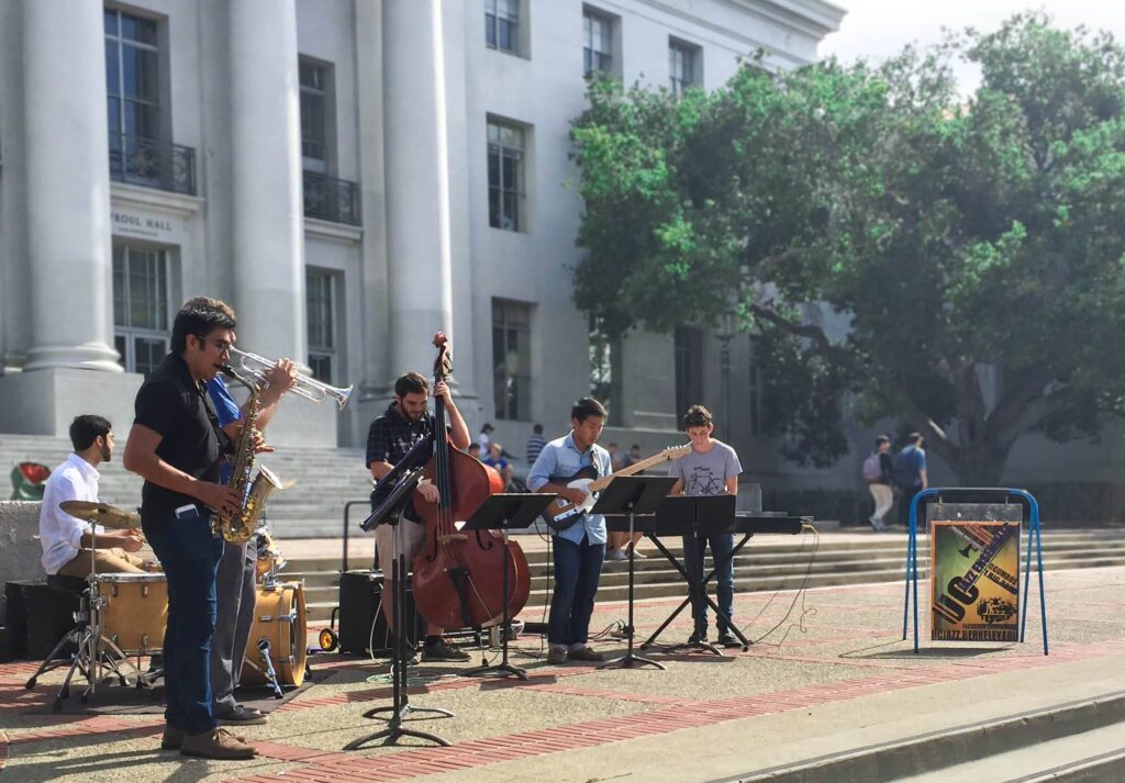 A group of five musicians performs on a sunny outdoor plaza. One plays the saxophone, another the double bass, with others on drums, keyboard, and electric guitar. Theyre in front of a large building with pillars and a tree in the background.