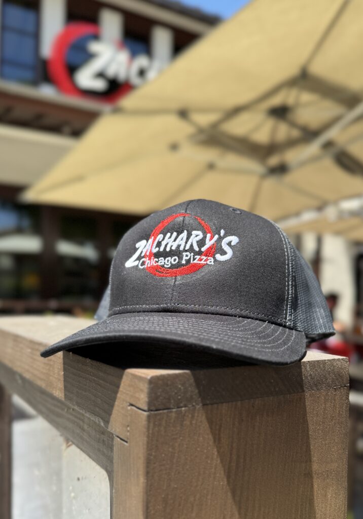 A black Zachary’s Chicago Pizza hat sits on a wooden railing outdoors, with a blurred restaurant and large beige patio umbrellas in the background.