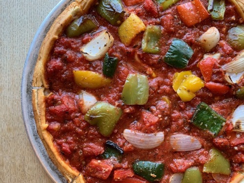 A close-up of a deep dish pizza topped with chunky tomato sauce, green and yellow bell peppers, onions, and pieces of tomato on a metal tray, shown on a light wooden surface.