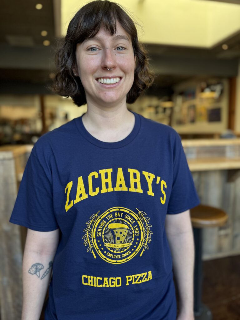 A smiling person with short brown hair stands indoors, wearing a navy blue Zacharys Chicago Pizza T-shirt with yellow lettering and a circular emblem in the center.