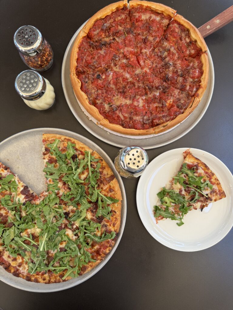 Two pizzas on a table: one deep dish with tomato sauce and cheese, and one thin crust topped with arugula. A slice of the thin crust pizza is served on a separate plate. Shakers with cheese and peppers are also on the table.