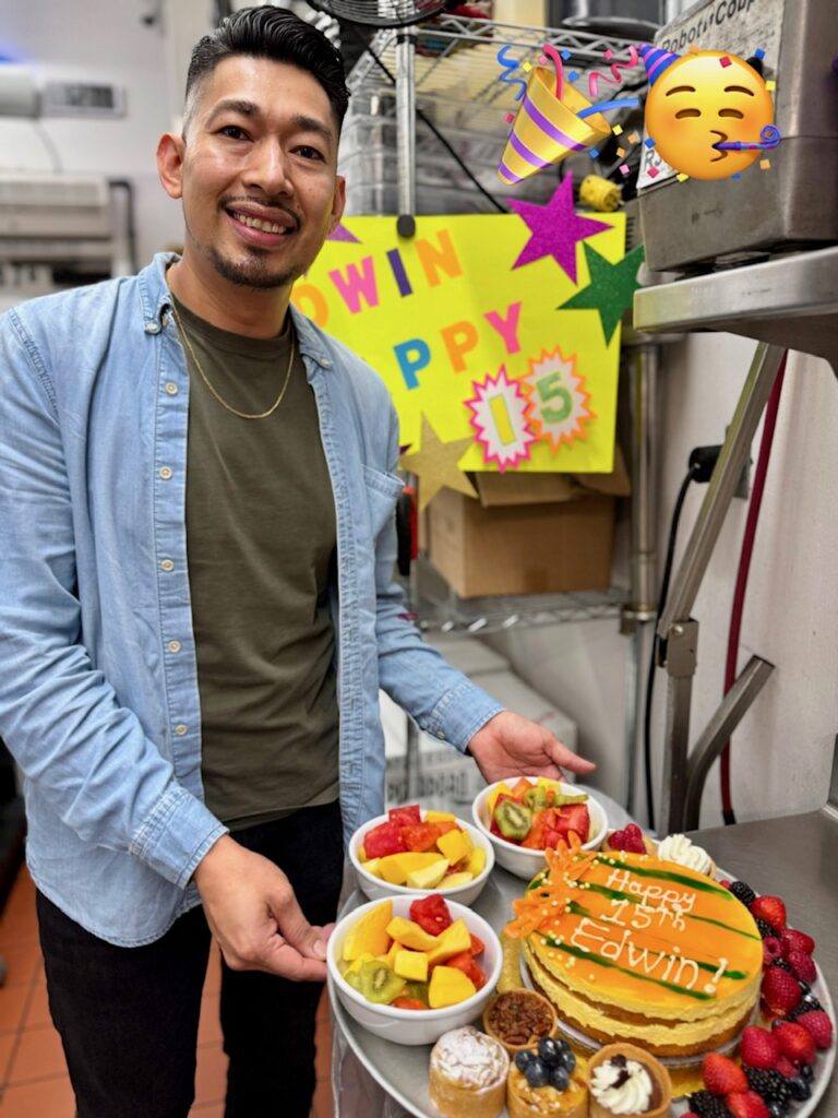 A man smiles while holding a tray with a Happy 15th Edwin cake and bowls of fruit. Behind him is a colorful sign reading WIN HAPPY 15. Party emojis decorate the top right corner of the image.