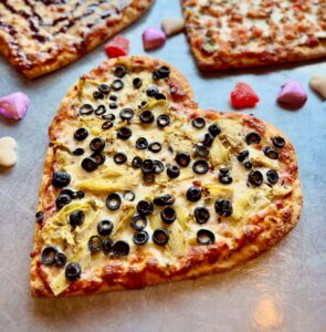 A heart-shaped pizza topped with black olives and artichokes sits on a table, surrounded by colorful candy hearts. Two other pizzas are partially visible in the background.