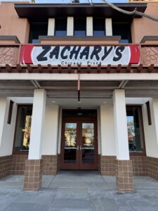 The entrance to Zacharys Chicago Pizza, featuring a large sign above the door with the restaurants name. The building has brown and white pillars and large windows on either side of the entrance.
