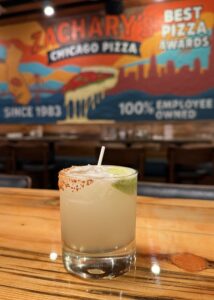 A margarita with a salted and chili rim and a lime wedge sits on a wooden table in a restaurant, with a colorful mural advertising Zacharys Chicago Pizza in the blurred background.