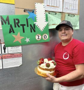 A man in a red shirt and cap holds a cake topped with strawberries. Behind him is a colorful sign with MARTIN and 20 in stars and an arrow, suggesting a celebration. Notices and papers are posted on the wall.