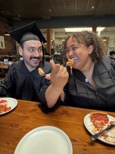 Two graduates in black caps and gowns smile and feed each other pizza slices while sitting at a wooden table in a restaurant, celebrating together with joy and laughter.