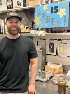 A bearded man in a black t-shirt and hat stands smiling in a kitchen. Behind him is a blue sign with stars that reads DARREN! 15. Trays of food, a jar of salsa, and bags of chips are on the counter.