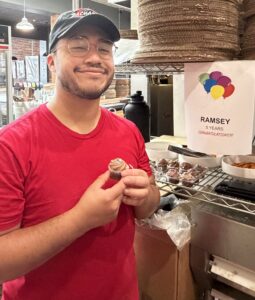 A smiling person in a red shirt and cap holds a cupcake in a kitchen. Behind them is a sign with colorful balloons that reads RAMSEY 5 YEARS CONGRATULATIONS! and a tray of more cupcakes.