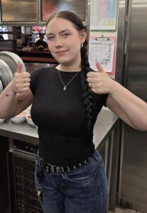 A woman with a long braid, wearing a black t-shirt and jeans, stands in a kitchen giving two thumbs up and smiling slightly at the camera.