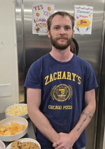 A man in a Zacharys Chicago Pizza shirt stands in front of celebratory signs, with bowls of popcorn, chips, and cookies on a table beside him.