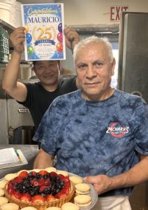 Two men in a kitchen celebrate 25 years of work. The man in front holds a fruit tart and cookies, while the man behind holds a colorful sign that says Congratulations Mauricio on 25 years of work.